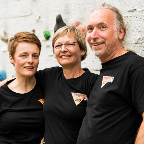 Drei Personen in schwarzen T-Shirts lächeln vor einer Kletterwand mit bunten Griffen.