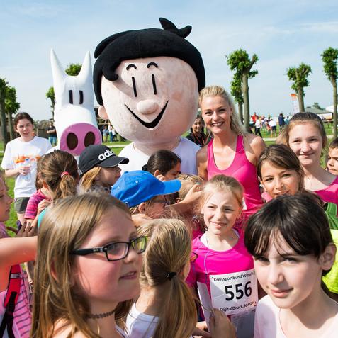Gruppenszene mit lächelnden Kindern und einer Frau in Sportkleidung neben einem großen Maskottchen auf einer Wiese.