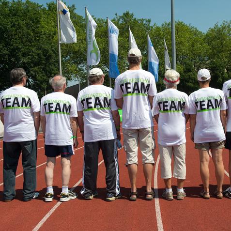Gruppe von sieben Personen in weißen T-Shirts mit dem Aufdruck "TEAM" auf einer Laufbahn, Blick von hinten.
