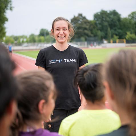 Lächelnde Trainerin auf der Laufbahn, umgeben von Kindern in Sportkleidung, die ihr gespannt zuhören.