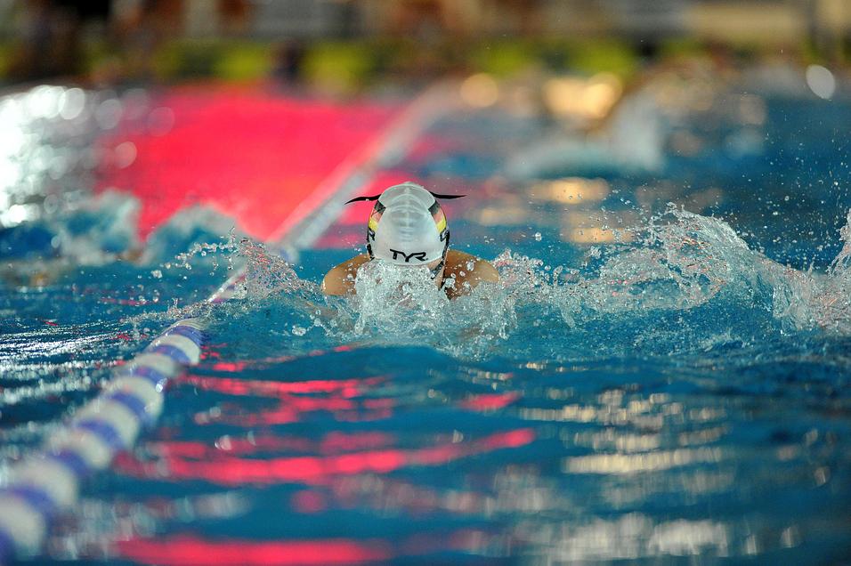 Schwimmerin mit weißer Badekappe taucht im Wasser, spritzt Wasser während des Schwimmens. Farbige Lichter reflektieren im Hintergrund.